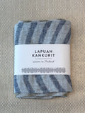 A folded woven textile with blue-gray and off-white striped pattern, wrapped in a white paper label that reads 'LAPUAN KANKURIT' and 'woven in Finland', placed on a textured beige surface.