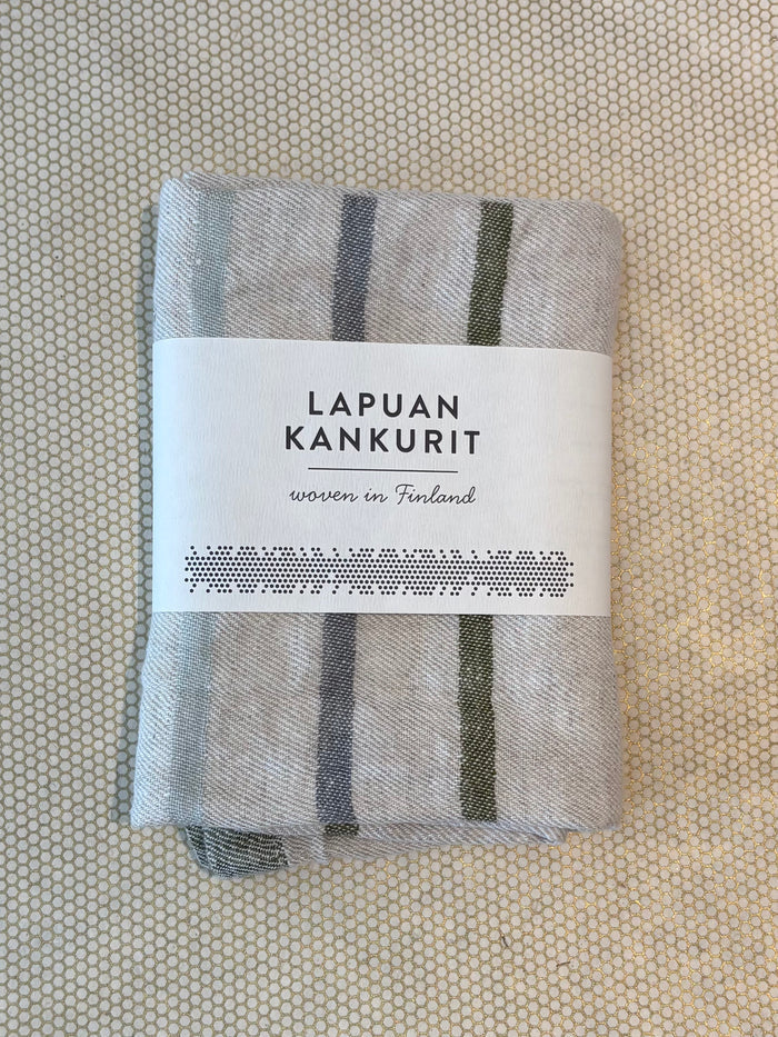 A folded light gray woven textile kitchen towel with dark gray vertical stripes, wrapped in a white paper label that reads 'LAPUAN KANKURIT' and 'woven in Finland,' placed on a textured beige surface.