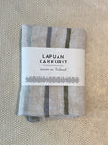 A folded light gray woven textile kitchen towel with dark gray vertical stripes, wrapped in a white paper label that reads 'LAPUAN KANKURIT' and 'woven in Finland,' placed on a textured beige surface.