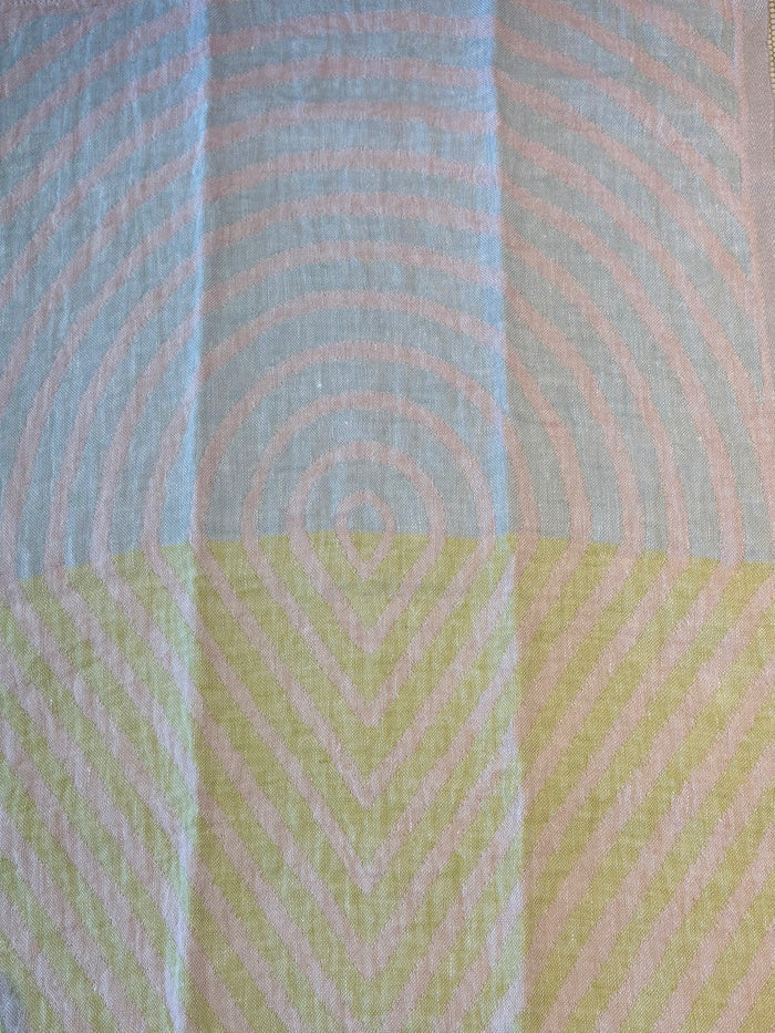 A close-up of a textured fabric with a geometric pattern, featuring concentric circles in light blue and pale pink on the top half, and chevron lines in pale yellow and pink on the bottom half.