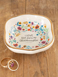 A ceramic trinket bowl with a gold rim and multicolor floral pattern, featuring the text 'I love that you’re my GRANDdaughter' on the bottom. The bowl is displayed on a wooden surface, and a ring is visible to the side.