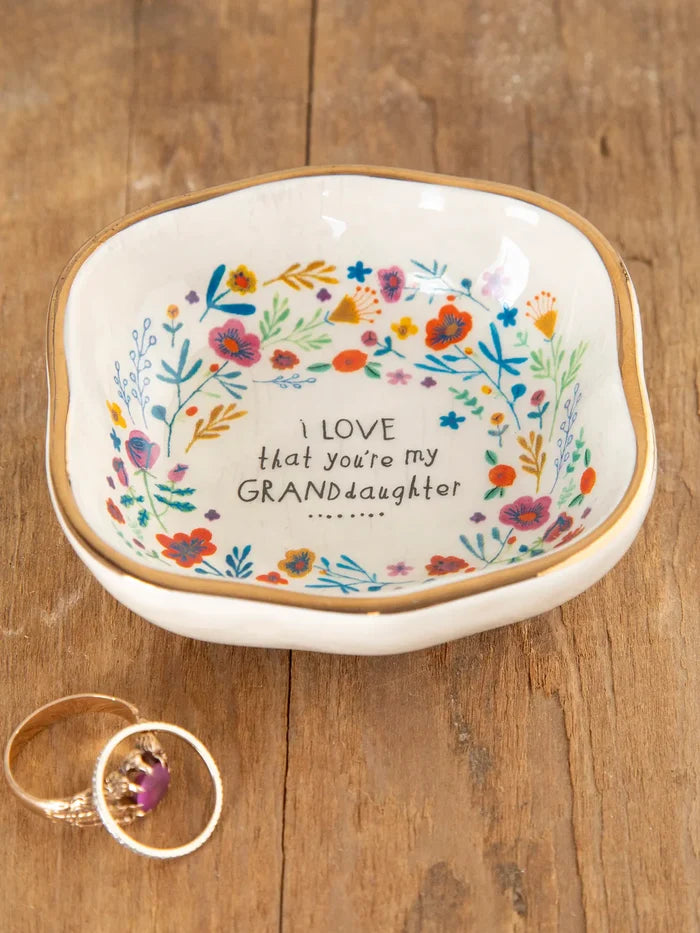 A ceramic trinket bowl with a gold rim and multicolor floral pattern, featuring the text 'I love that you’re my GRANDdaughter' on the bottom. The bowl is displayed on a wooden surface, and a ring is visible to the side.