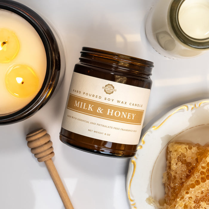 Rustic Strength, Candle, 8oz, Amber Glass photoshoot image taken from above of candle jar on it's side, with a lit, 16oz candle beside it, a glass pitcher of milk and a plate with blocks of honey comb with a wooden honey utensil