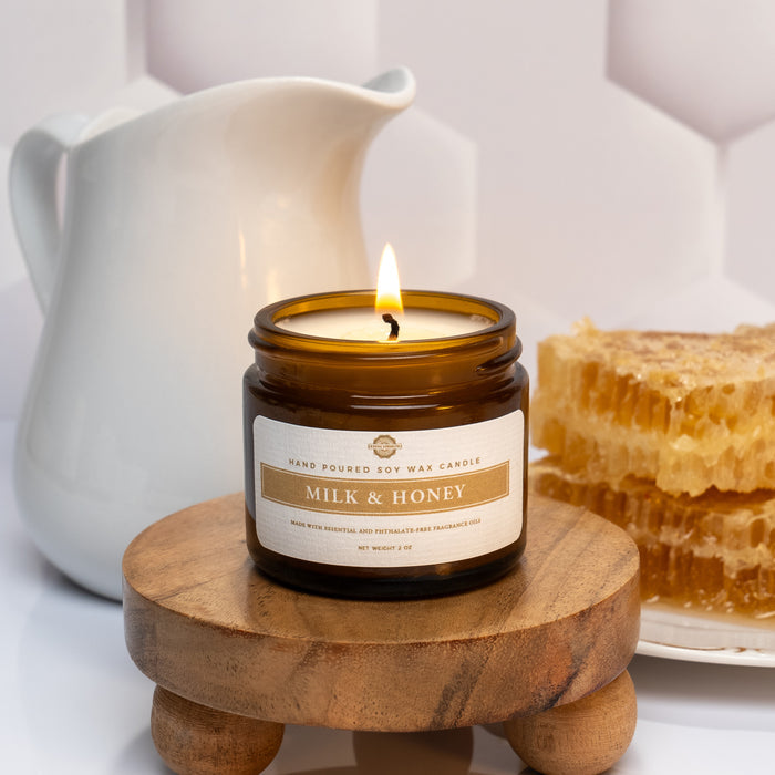 Rustic Strength, Candle, 2oz, Amber Glass photoshoot image of lit candle set on a wooden platform with a white honeycomb texture background, set in front of a white ceramic pitcher and a plate with blocks of honey comb