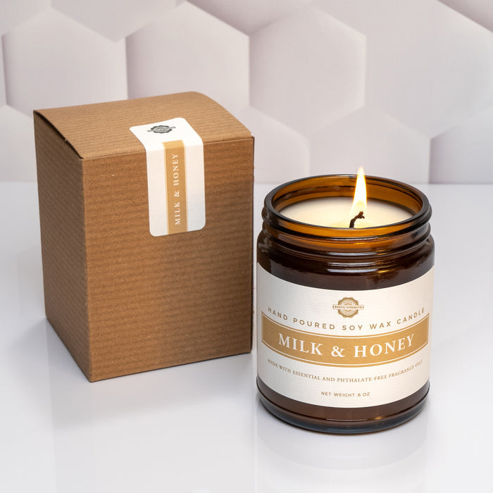 Rustic Strength, Candle, 8oz, Amber Glass Milk & Honey, photoshoot image of lit candle with a white honeycomb texture background, set in front of the box that the candle comes in.