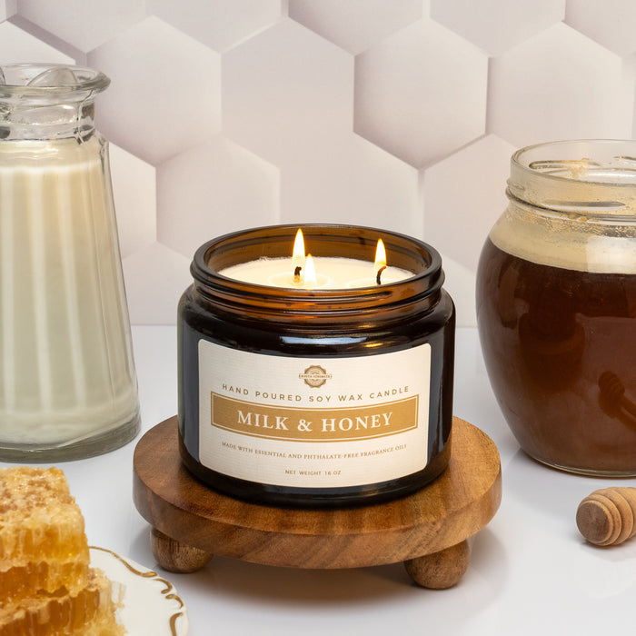 Rustic Strength, Candle, 16oz, Amber Glass with 3 wicks, Milk & Honey, photoshoot image of lit candle set on a wooden platform with a white honeycomb texture background, set in front of a glass pitcher filled with milk, a glass jar filled with honey and a plate with blocks of honey comb