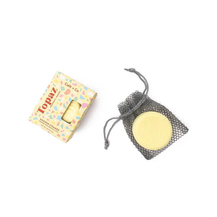 A shower steamer in the form of a yellow rounded object next to its open, decorative box with multicolored design and a mesh bag.
