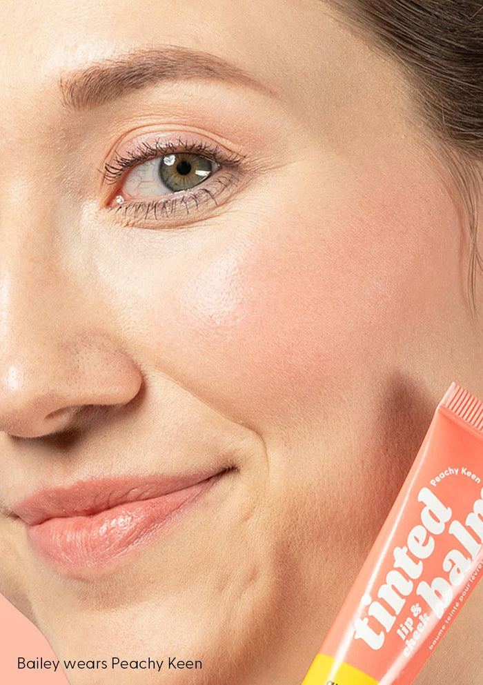 Close-up of a person's face with a tube of tinted balm labeled 'Peachy Keen'.