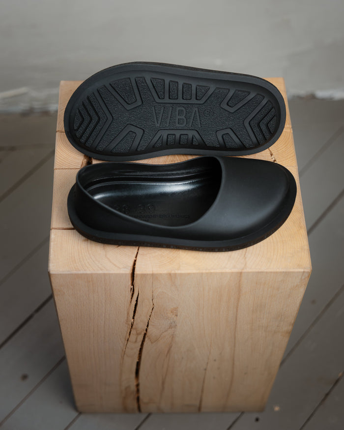 Black slip-on clog with textured rubber sole displayed on natural wood block.