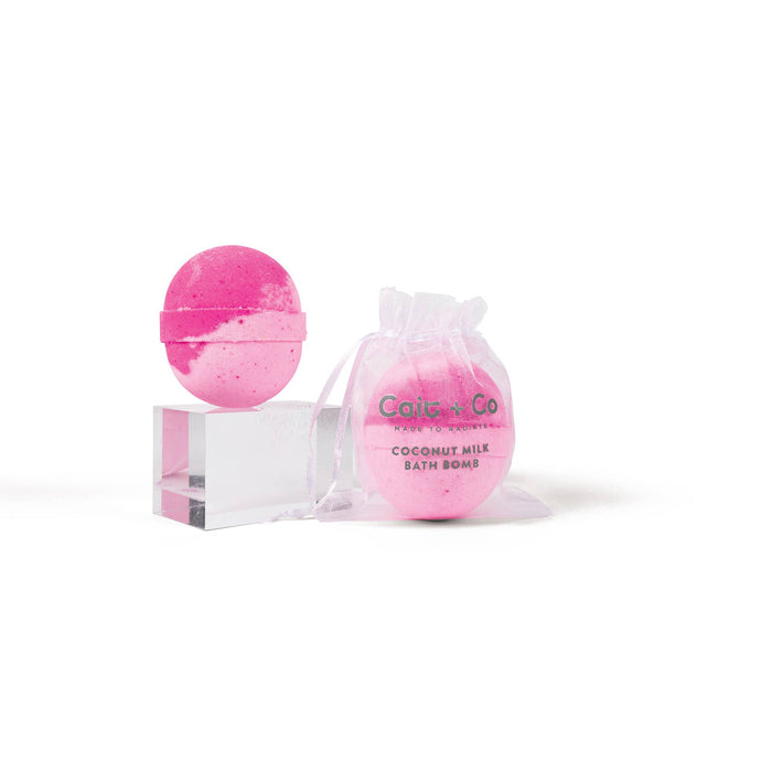 A pink bath bomb with a clear packaging that reads 'CAIT + CO. BATH BOMB' and a decorative bow, placed on a white stand against a white background.