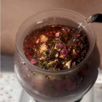 Hot water being poured into a glass teapot filled with loose leaf tea.